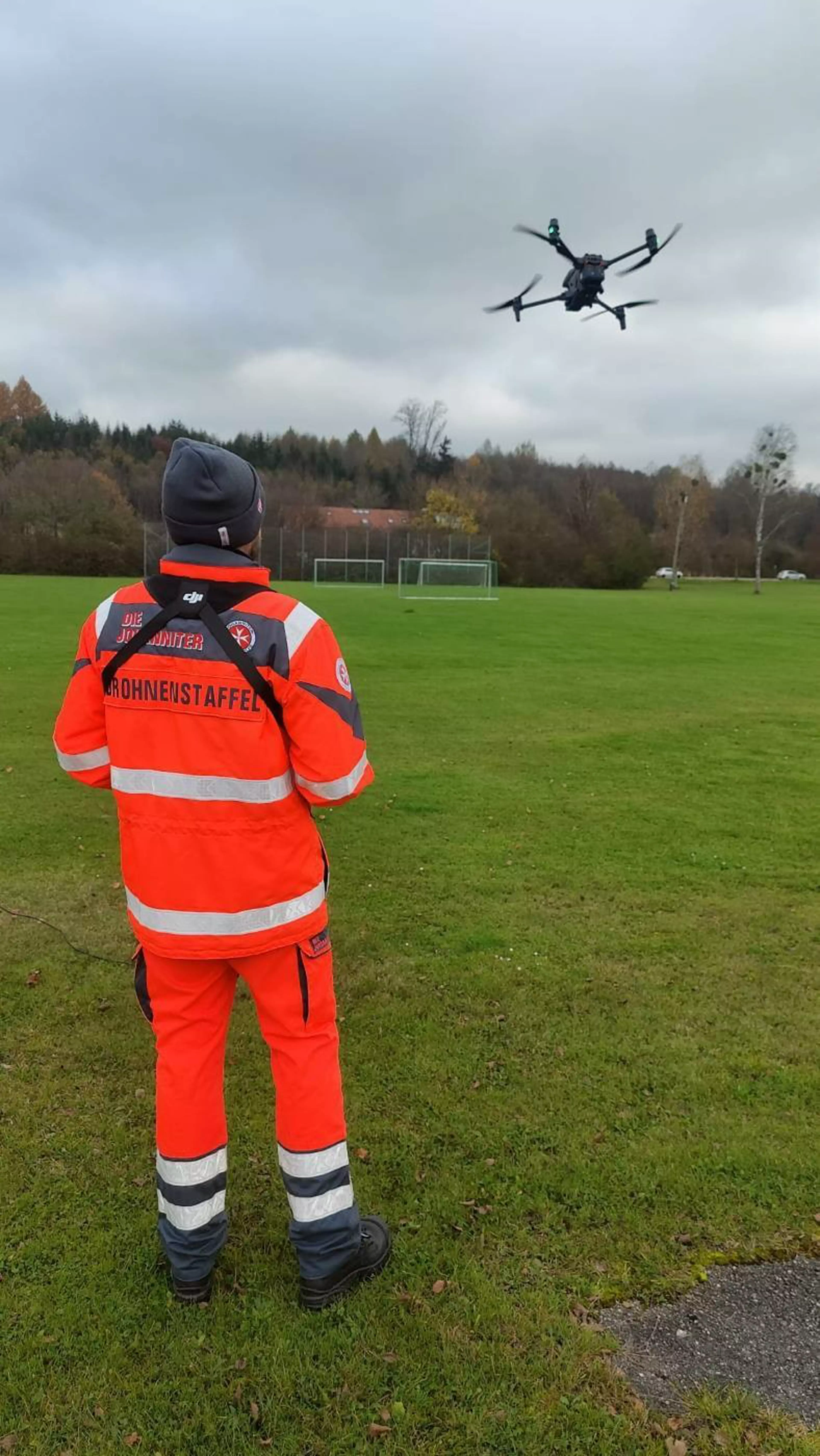 Ein Mitglied der Johanniter-Drohnenstaffel steht in leuchtend orangefarbener Einsatzkleidung mit Reflexstreifen auf einem grünen Rasenplatz. Die Person trägt eine dunkle Mütze und steuert eine schwarze Quadrocopter-Drohne, die in der Luft über einem Fußballfeld schwebt. Im Hintergrund sind bewaldete Hügel unter einem bewölkten Himmel zu sehen.