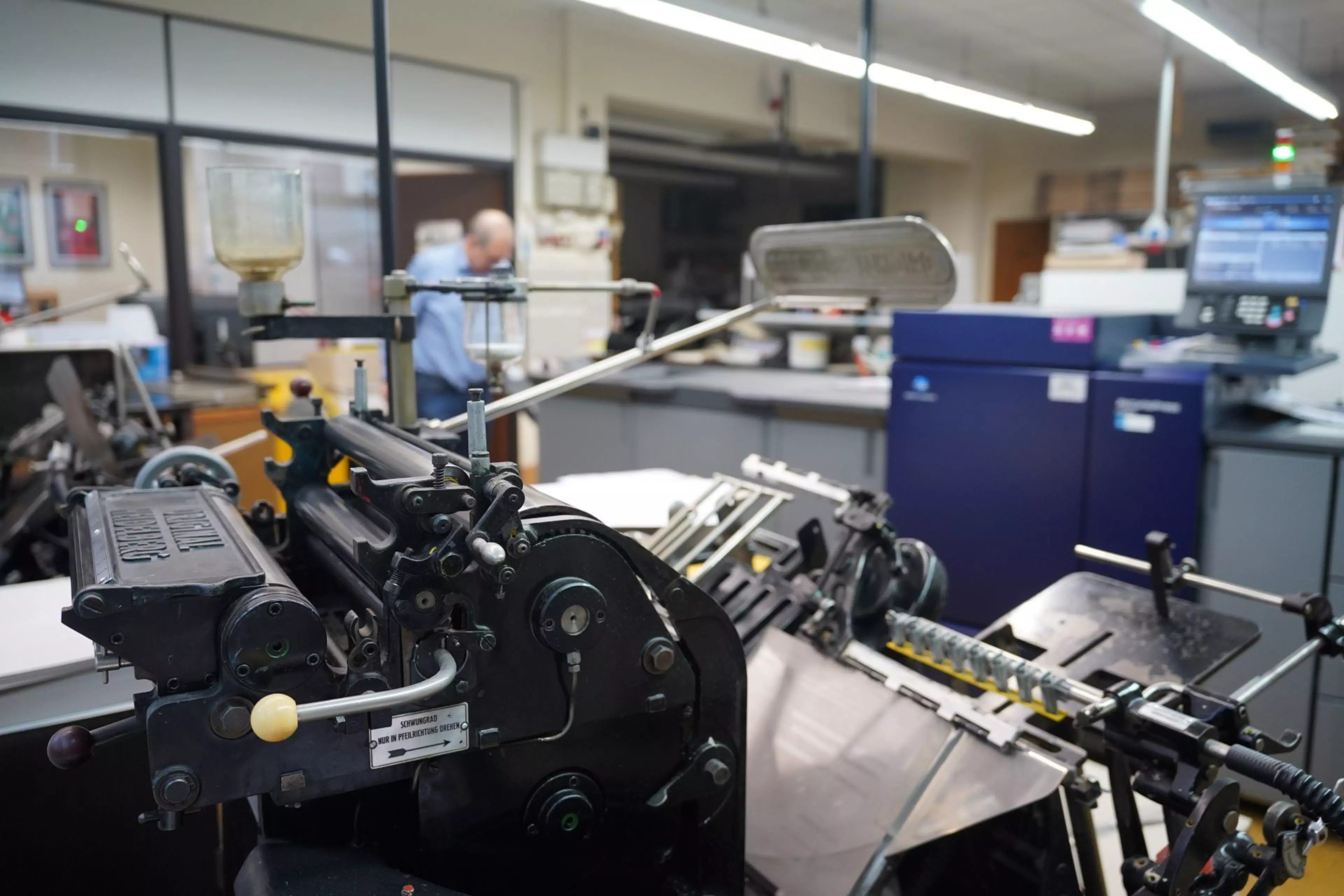 Historische Druckmaschine in einer modernen Druckerei, im Hintergrund arbeitet eine Person an weiteren Produktionssystemen.