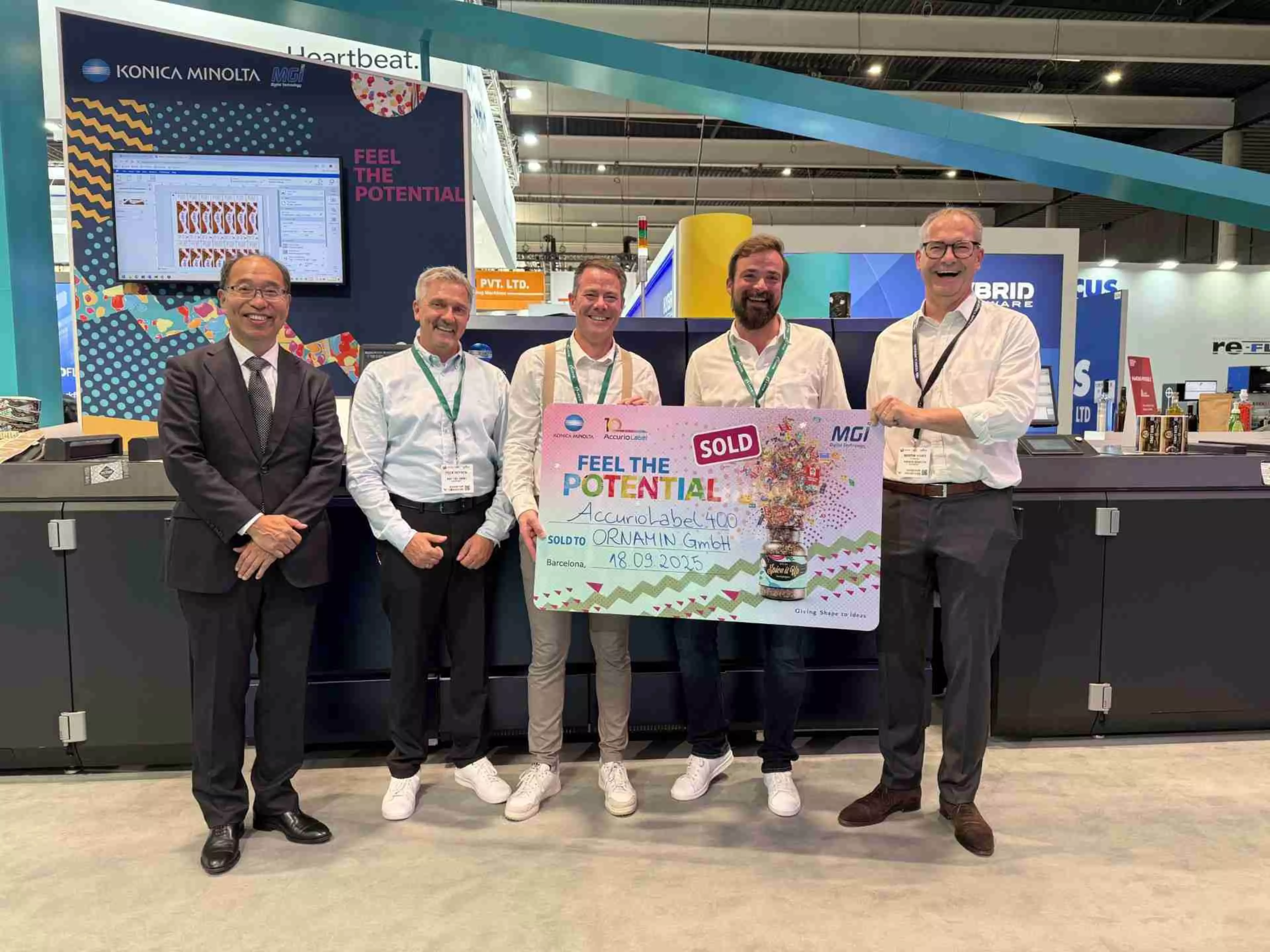 Fünf lächelnde Männer posieren auf dem Messestand von Konica Minolta auf der Labelexpo in Barcelona. Sie halten eine Plakette, die den Verkauf der AccurioLabel 400 an die Ornamin GmbH feiert.
