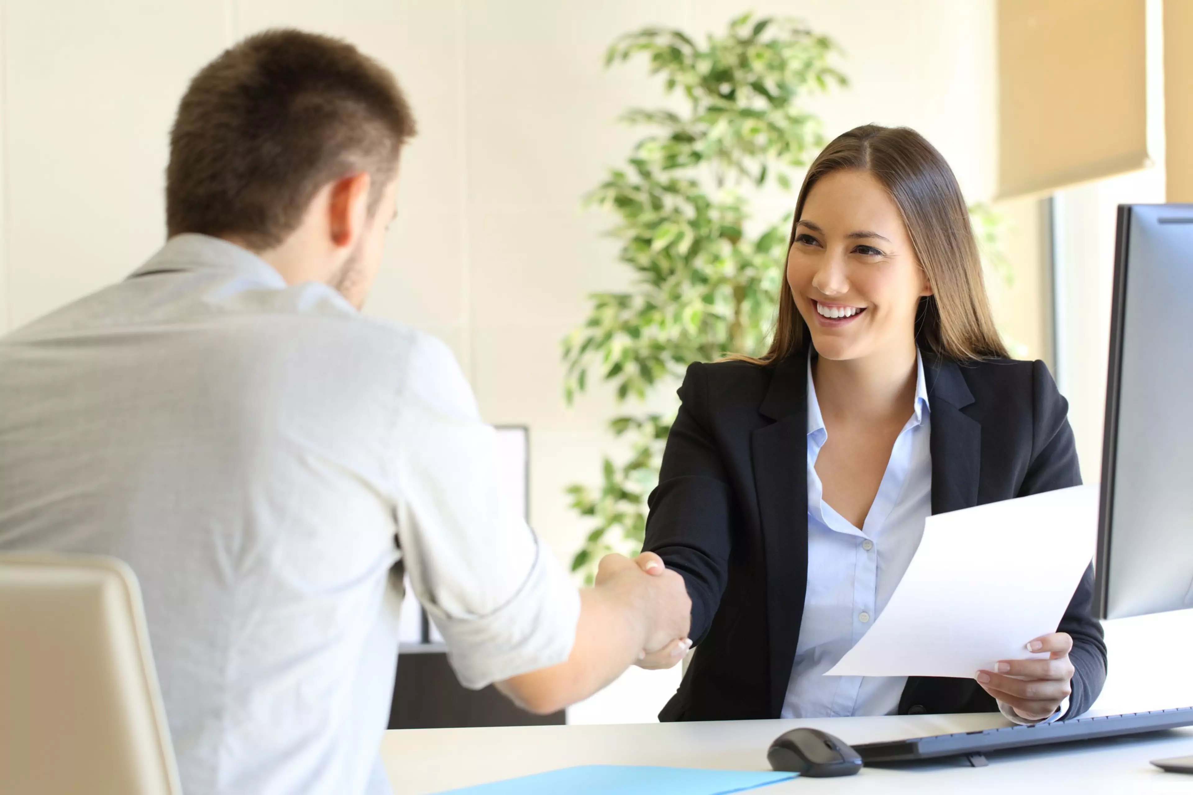 Frau im Business-Outfit begrüßt einen Bewerber mit einem Handschlag in einem modernen Büro – beide lächeln, die Stimmung ist positiv und professionell.