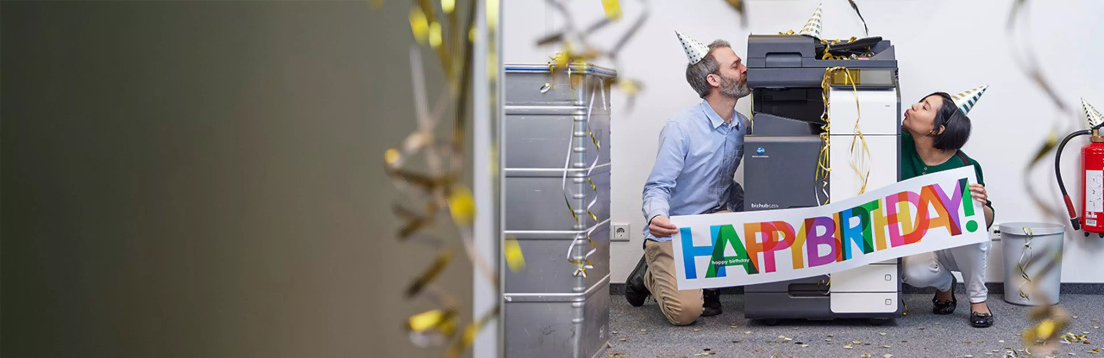Two people wearing party hats celebrate next to a copier with a “Happy Birthday” sign.