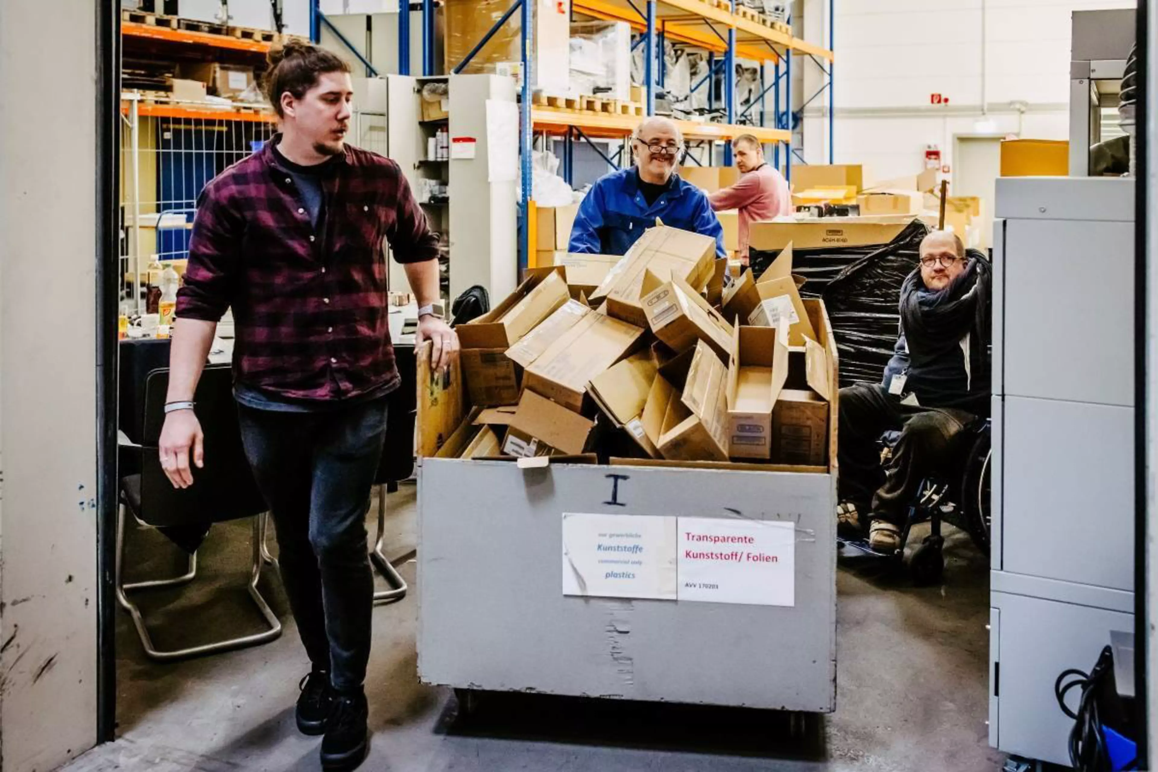 Gemini hat gesagt
In einem Logistiklager schiebt ein Mann im karierten Hemd einen großen Rollcontainer voller flachgelegter Kartons. Ein lächelnder Mitarbeiter in Arbeitskleidung geht dahinter, während im Hintergrund Regale und weitere Personen zu sehen sind.