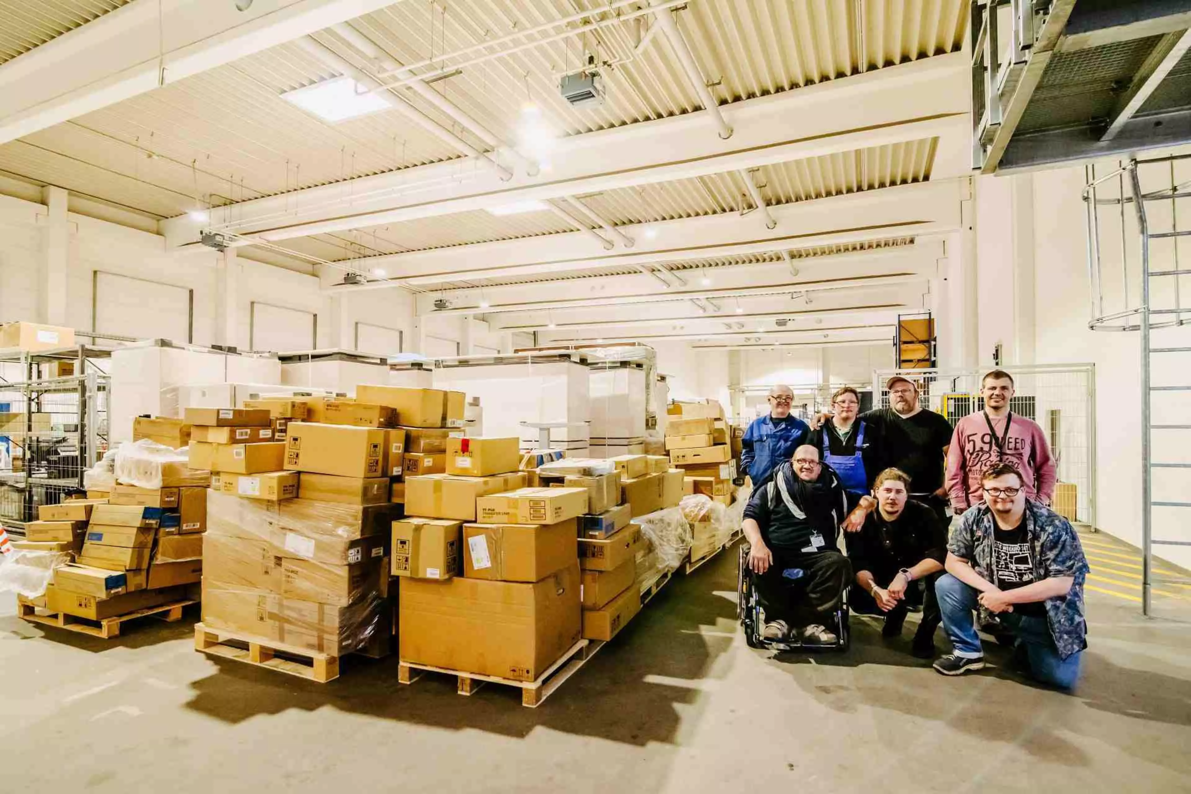 In einer großen Lagerhalle posiert eine Gruppe von acht Mitarbeitern, darunter ein Mann im Rollstuhl, vor aufgeschichteten Kartonstapeln auf Paletten. Einige stehen, andere knien. Der Hintergrund zeigt Regale und die industrielle Struktur der Halle.