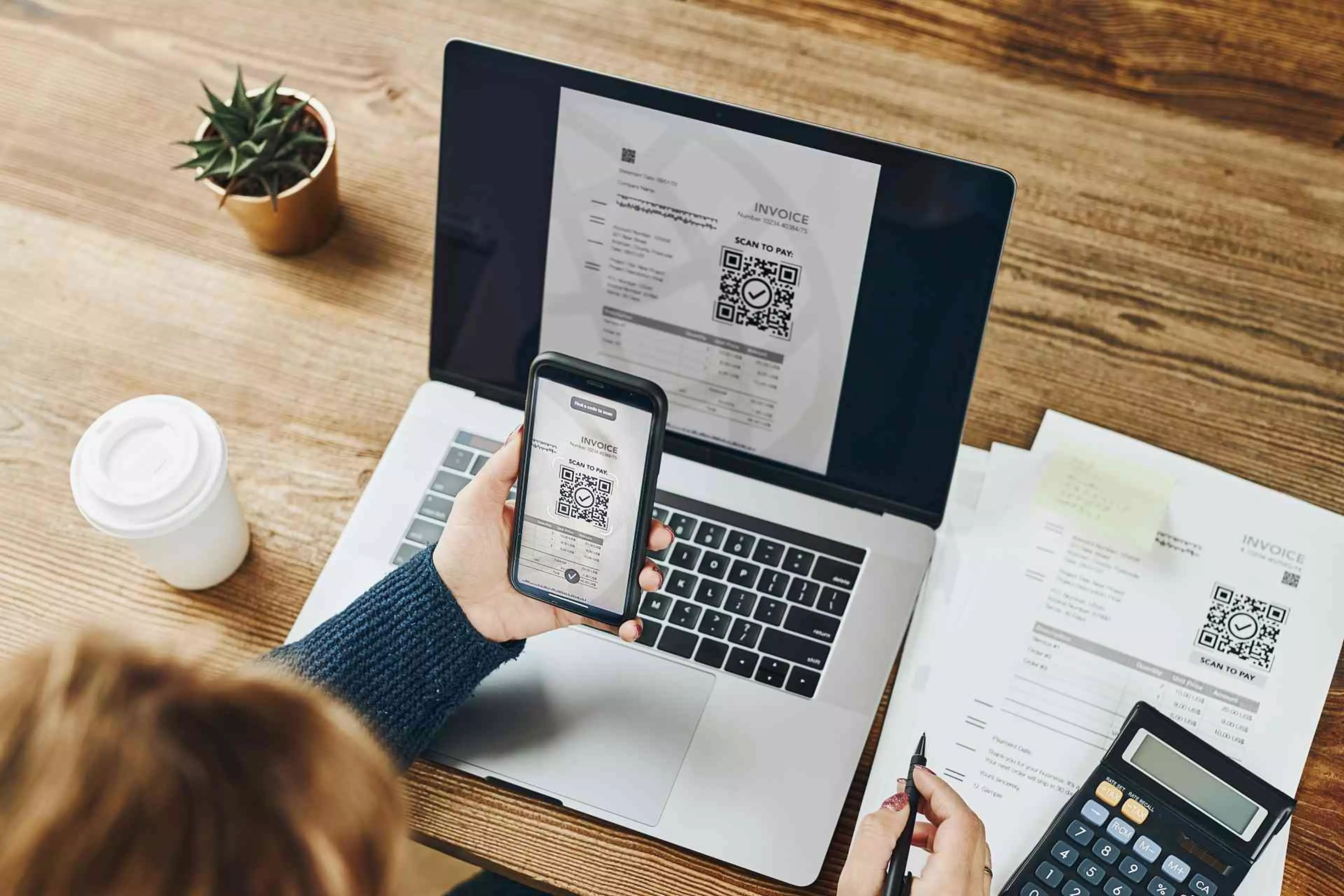 Person scannt mit ihrem Smartphone einen QR-Code auf einer Rechnung, die auf einem Laptopbildschirm angezeigt wird. Auf dem Tisch liegen weitere Rechnungen und ein Taschenrechner.
