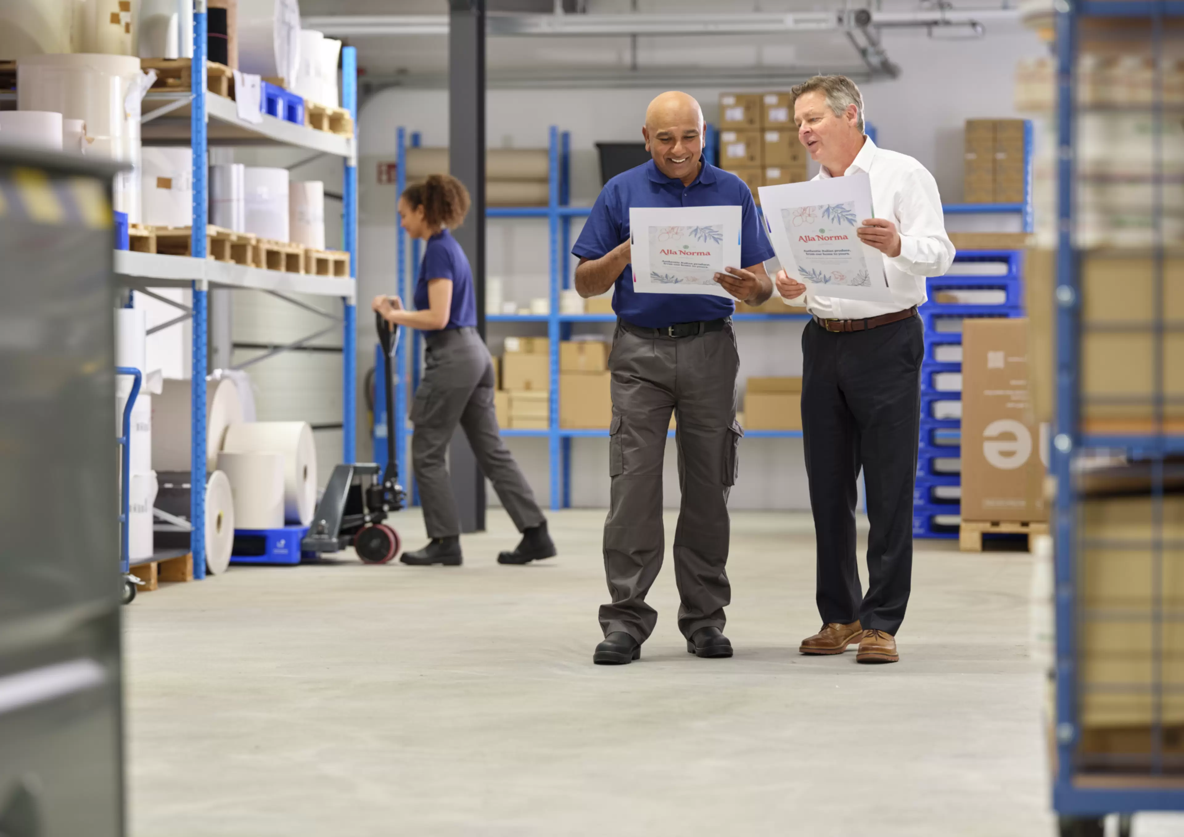 Two printing industry professionals reviewing printed marketing materials in a warehouse, with a colleague handling paper rolls and printing supplies stored on shelves in the background.