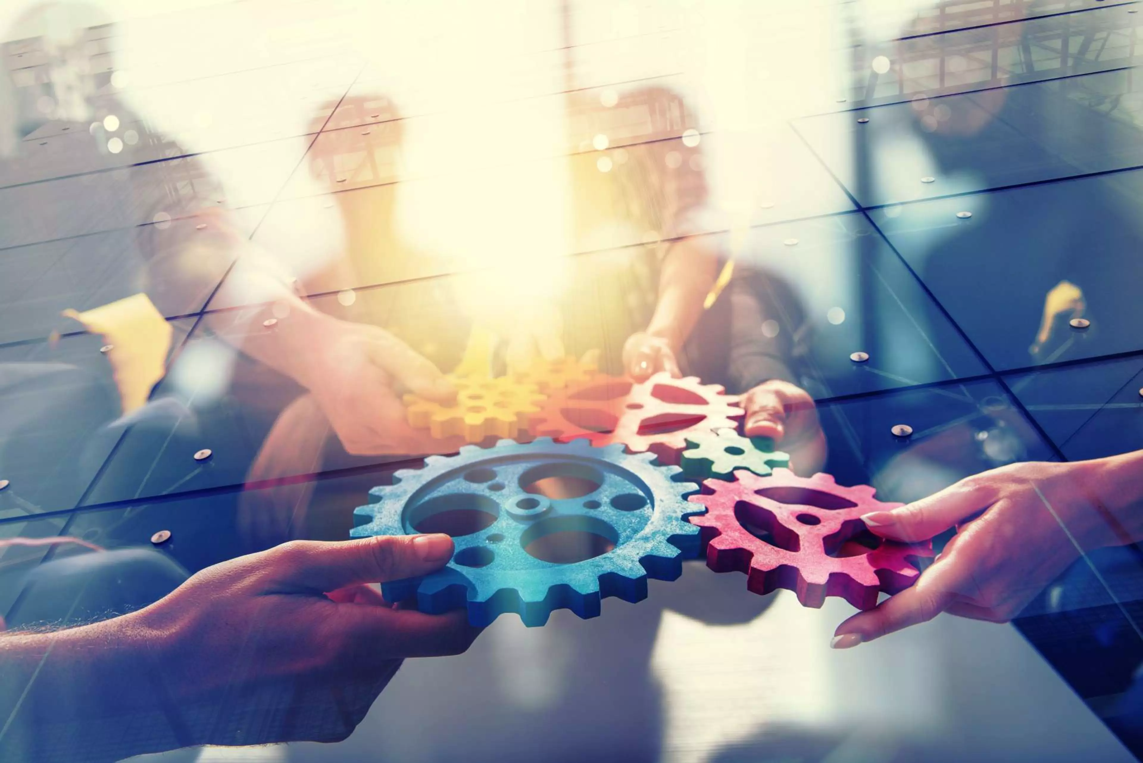 Mehrere Hände greifen nach bunten Zahnrädern, die symbolisch für Zusammenarbeit und Teamarbeit stehen. Die Aufnahme vermittelt das Konzept von Innovation und partnerschaftlicher Zusammenarbeit.