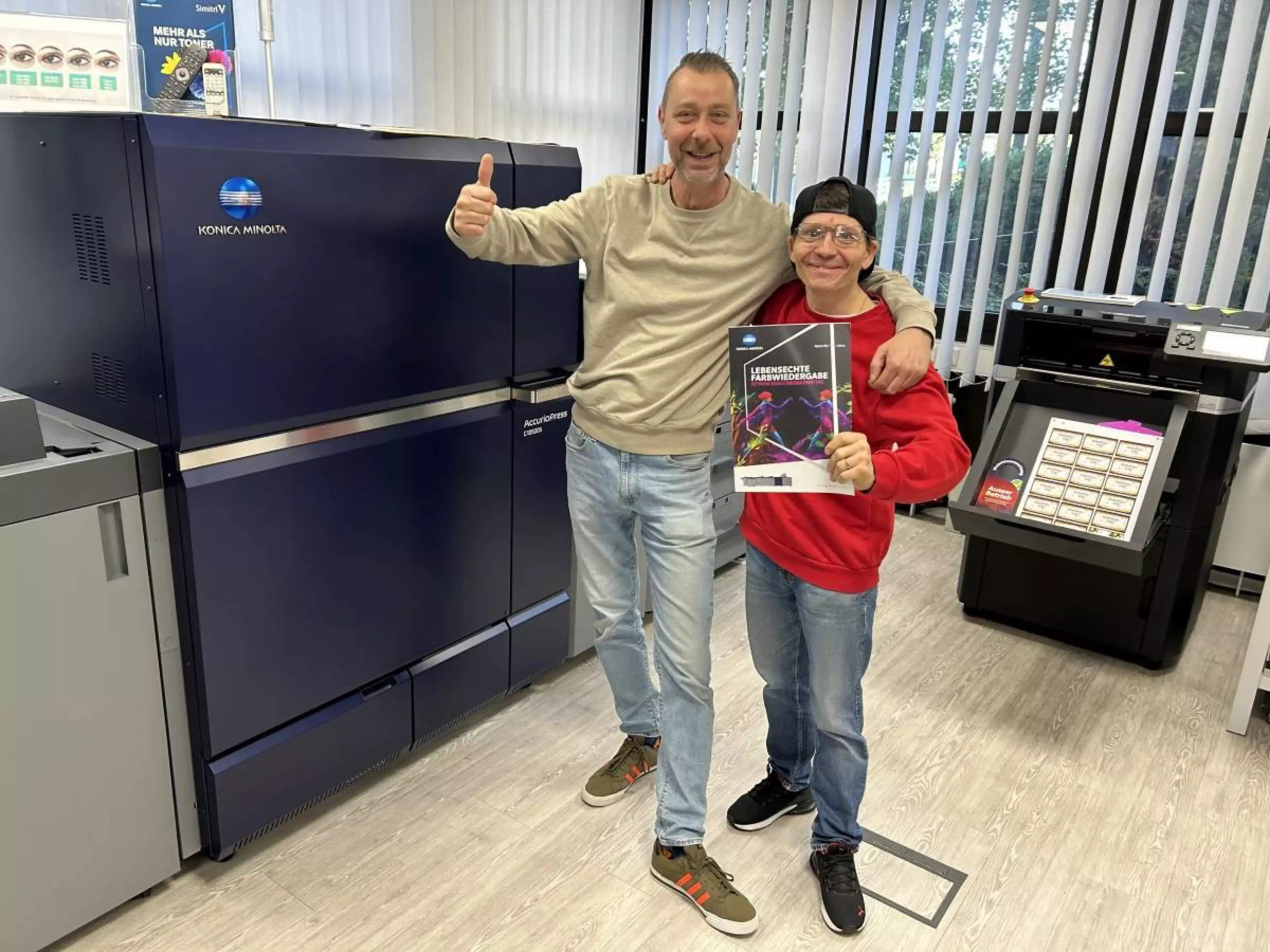 Zwei Männer stehen freudig in einem hellen Druckraum vor einer großen, dunkelblauen Konica Minolta Industrie-Druckmaschine. Der linke Mann trägt ein beiges Sweatshirt, helle Jeans und gibt ein Daumen-hoch-Zeichen. Der rechte Mann im roten Pullover hält eine Broschüre mit dem Titel „Lebensechte Farbwiedergabe“ in der Hand. Beide lächeln in die Kamera und haben kollegial die Arme umeinander gelegt.
