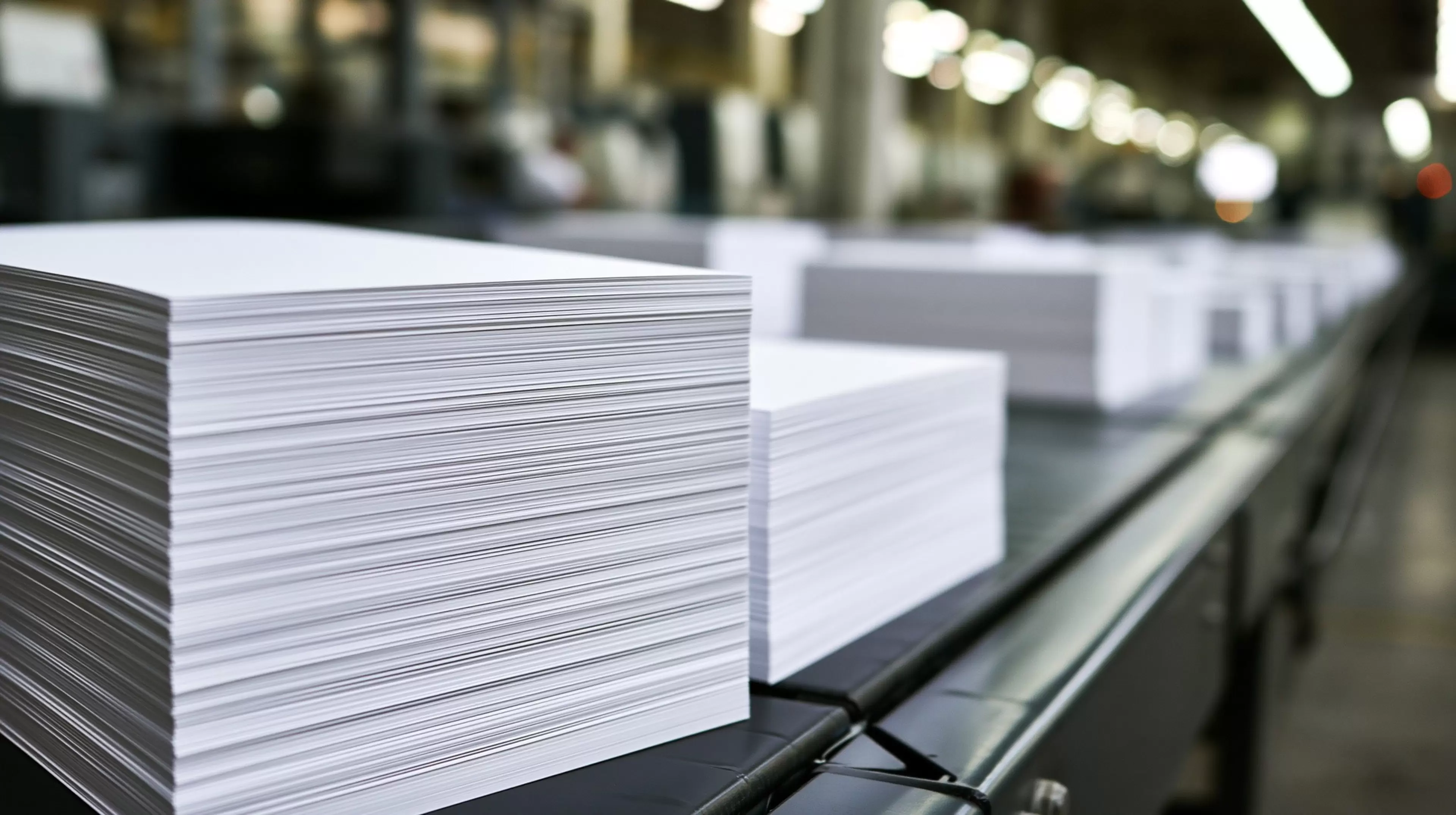 A sharp, eye-level close-up captures a long conveyor belt in a bustling industrial printing facility. Two prominent, neatly stacked piles of freshly printed white paper sit in the foreground, with several blurred stacks fading into the background. The scene is bathed in bright, professional warehouse lighting, highlighting the crisp edges of the paper. The depth of field is shallow, emphasizing the texture of the stacked paper. --ar 16:9 --profile kni3ml9 --hd --v 8.2 Job ID: 235c5e99-abe5-4de2-86ff-6df9124129d3