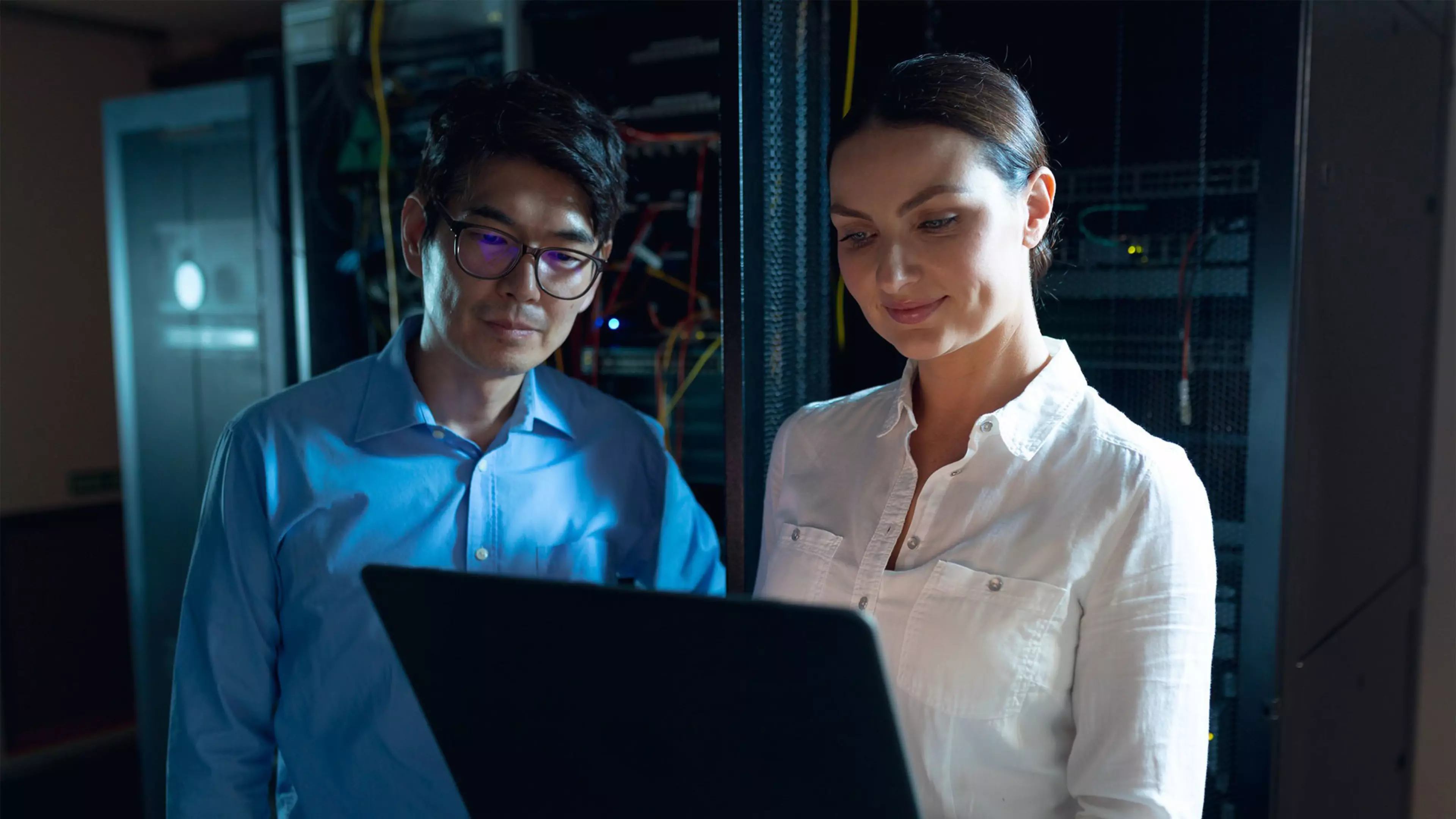 Un hombre y una mujer en una sala de servidores trabajan frente a un ordenador portátil