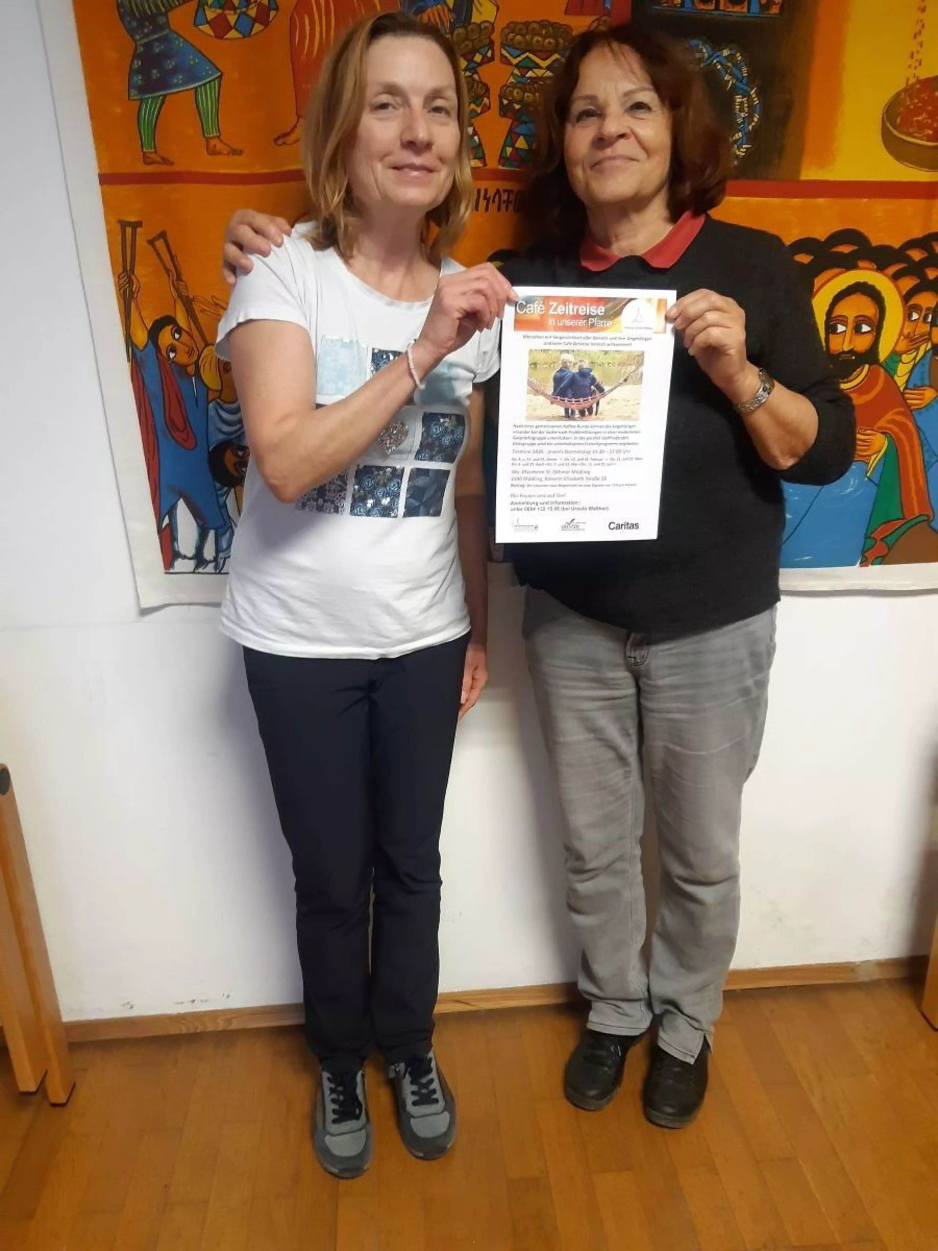 Zwei Frauen stehen lächelnd nebeneinander vor einer bunten Wandmalerei und halten gemeinsam ein Informationsblatt mit der Überschrift „Café Zeitreise“ in die Kamera. Die linke Frau trägt ein weißes T-Shirt und dunkle Hosen, die rechte Frau einen schwarzen Pullover und graue Jeans. Das Plakat enthält Informationen über ein Treffen für Menschen mit Vergesslichkeit und deren Angehörige in einer Pfarre.