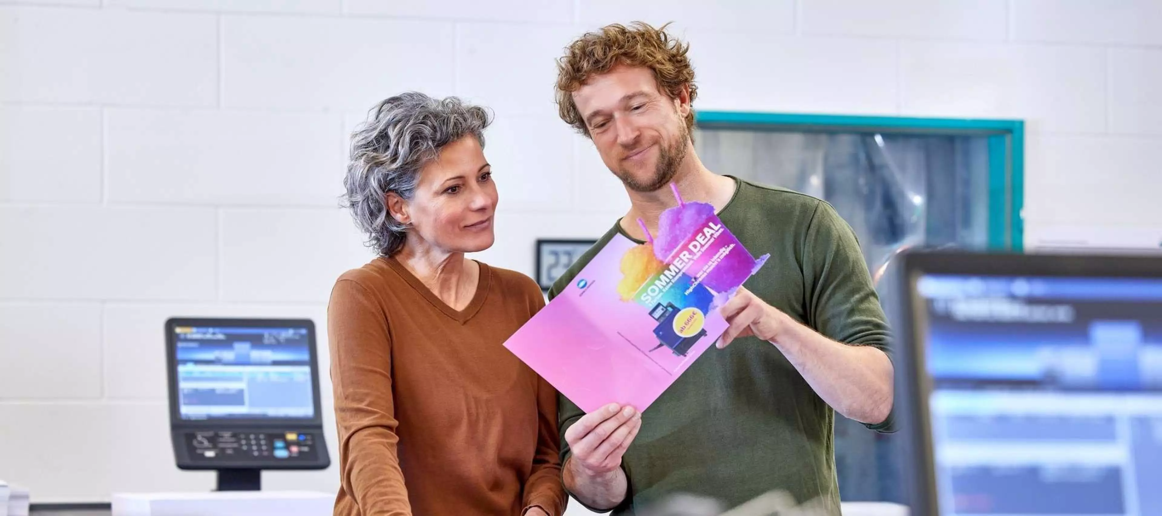 Das Bild zeigt zwei Personen, die in einem Druckraum stehen und eine gedruckte Broschüre oder ein Plakat betrachten. Beide scheinen mit der gedruckten Qualität zufrieden zu sein. Die Person links trägt ein braunes Oberteil, die Person rechts ein grünes. Im Hintergrund ist eine Druckmaschine sichtbar, die den Eindruck eines modernen Produktionsdrucks erweckt.