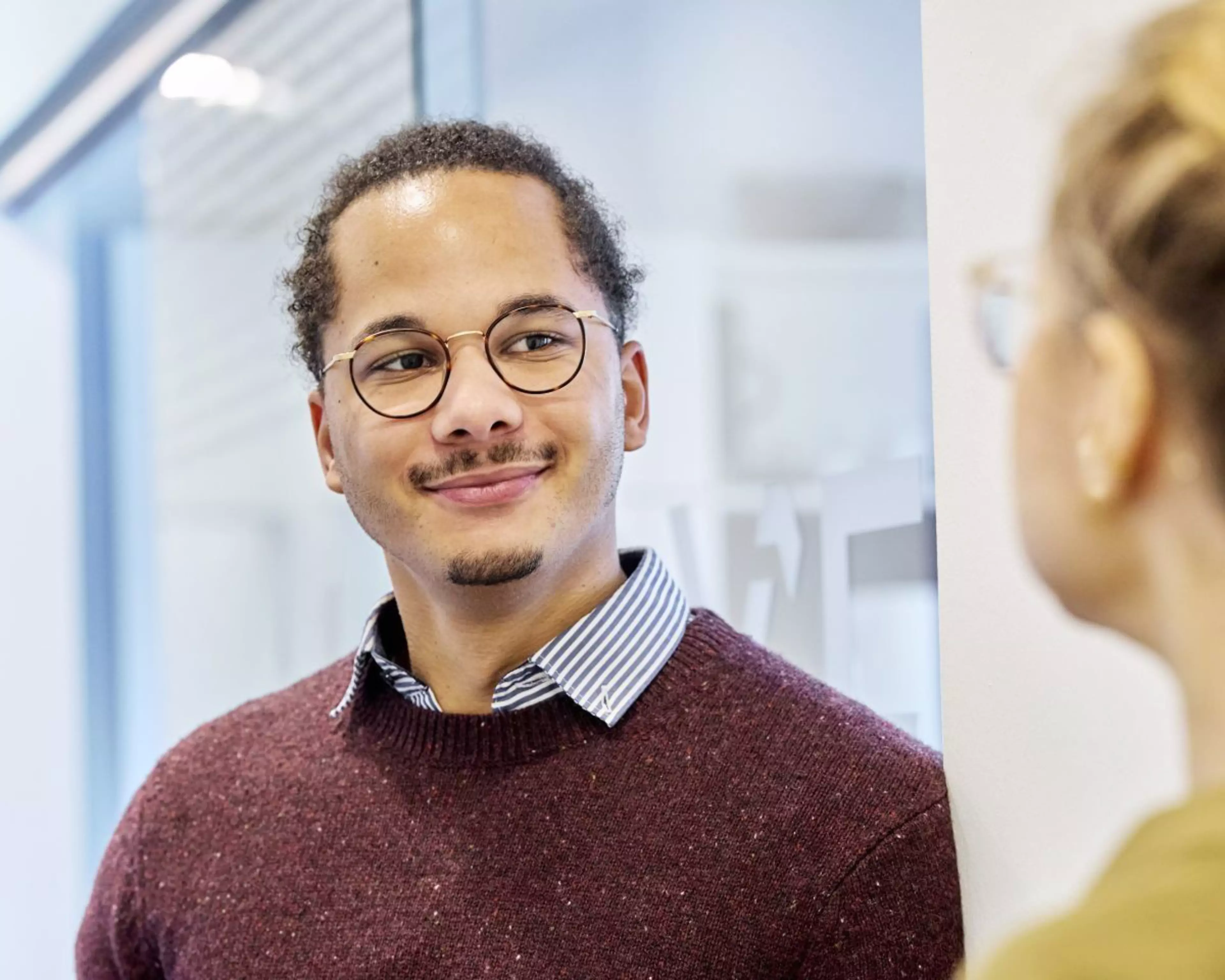 Marvin Sobottka lächelt freundlich in einer Büroumgebung; er trägt eine Brille, einen weinroten Strickpullover und ein gestreiftes Hemd.
