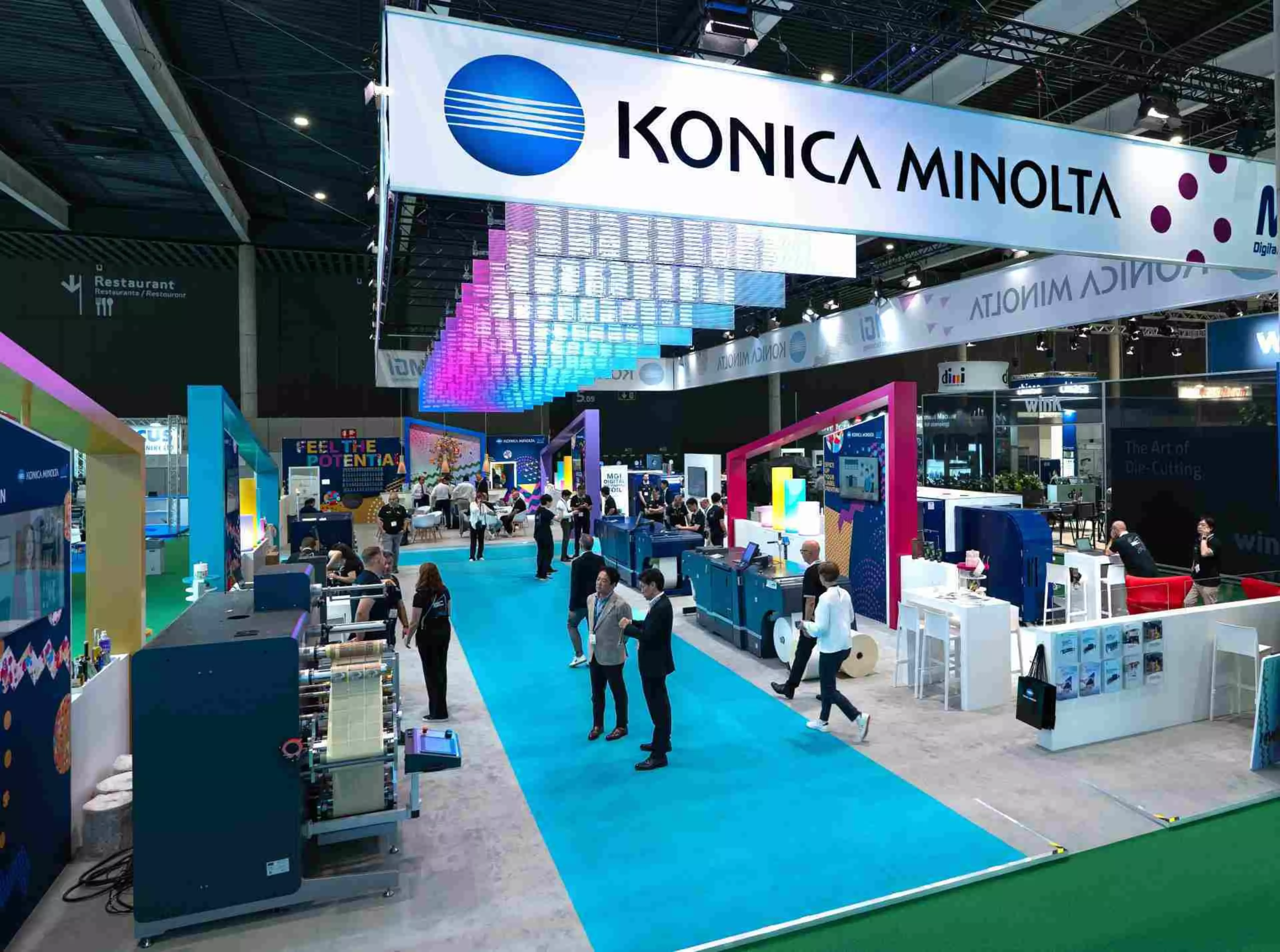 Das Bild zeigt den belebten Messestand von Konica Minolta auf der Labelexpo in Barcelona. Ein großes Banner mit dem Firmennamen hängt über einem markanten blauen Teppich, der durch den Stand führt, auf dem sich zahlreiche Besucher und Mitarbeiter befinden.