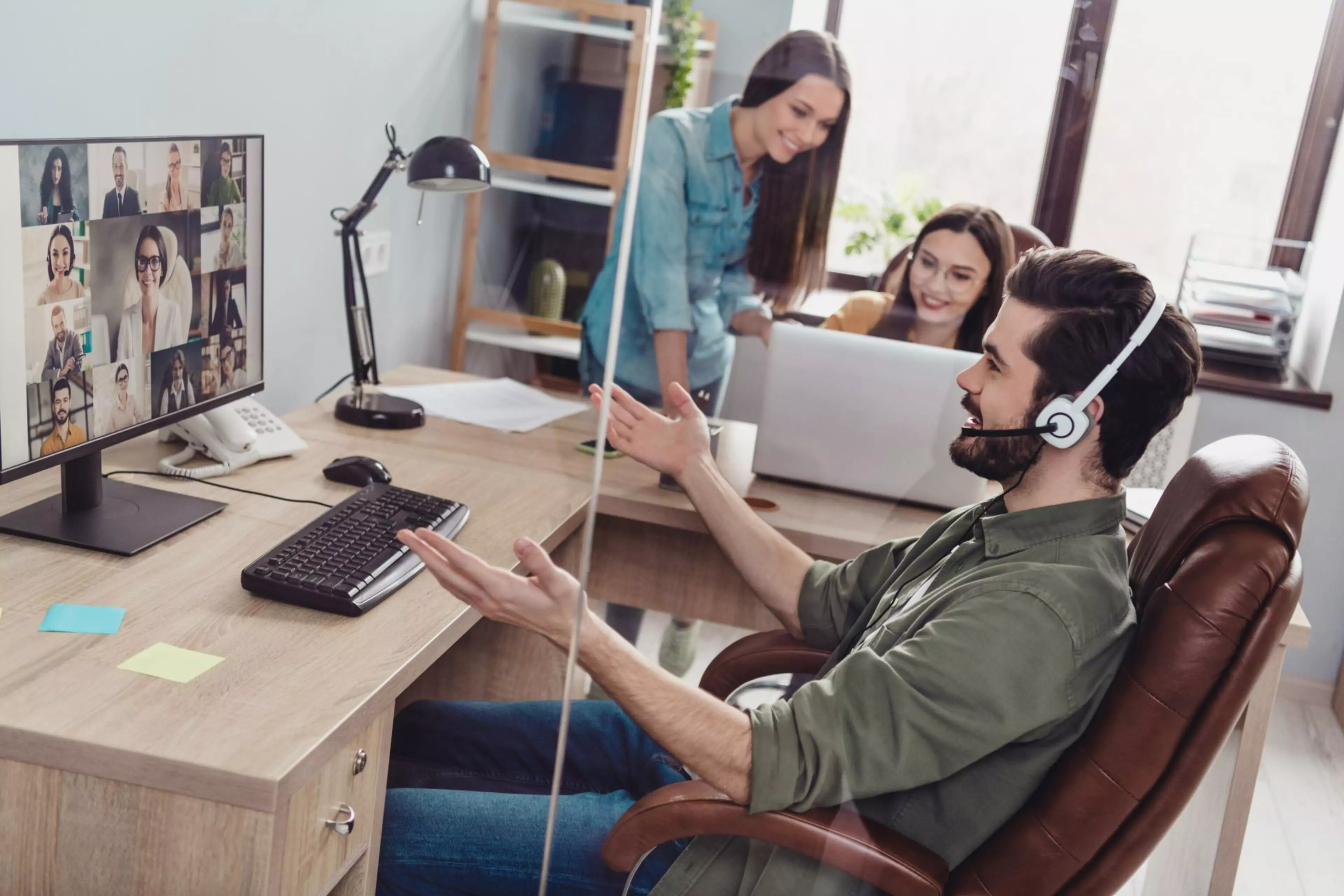 Junger Mann mit Headset im Video-Call an einem Schreibtisch, auf dem Bildschirm sind mehrere Teilnehmende zu sehen; im Hintergrund zwei Kolleginnen an einem Laptop – Szene aus einem modernen, hybriden Arbeitsumfeld.