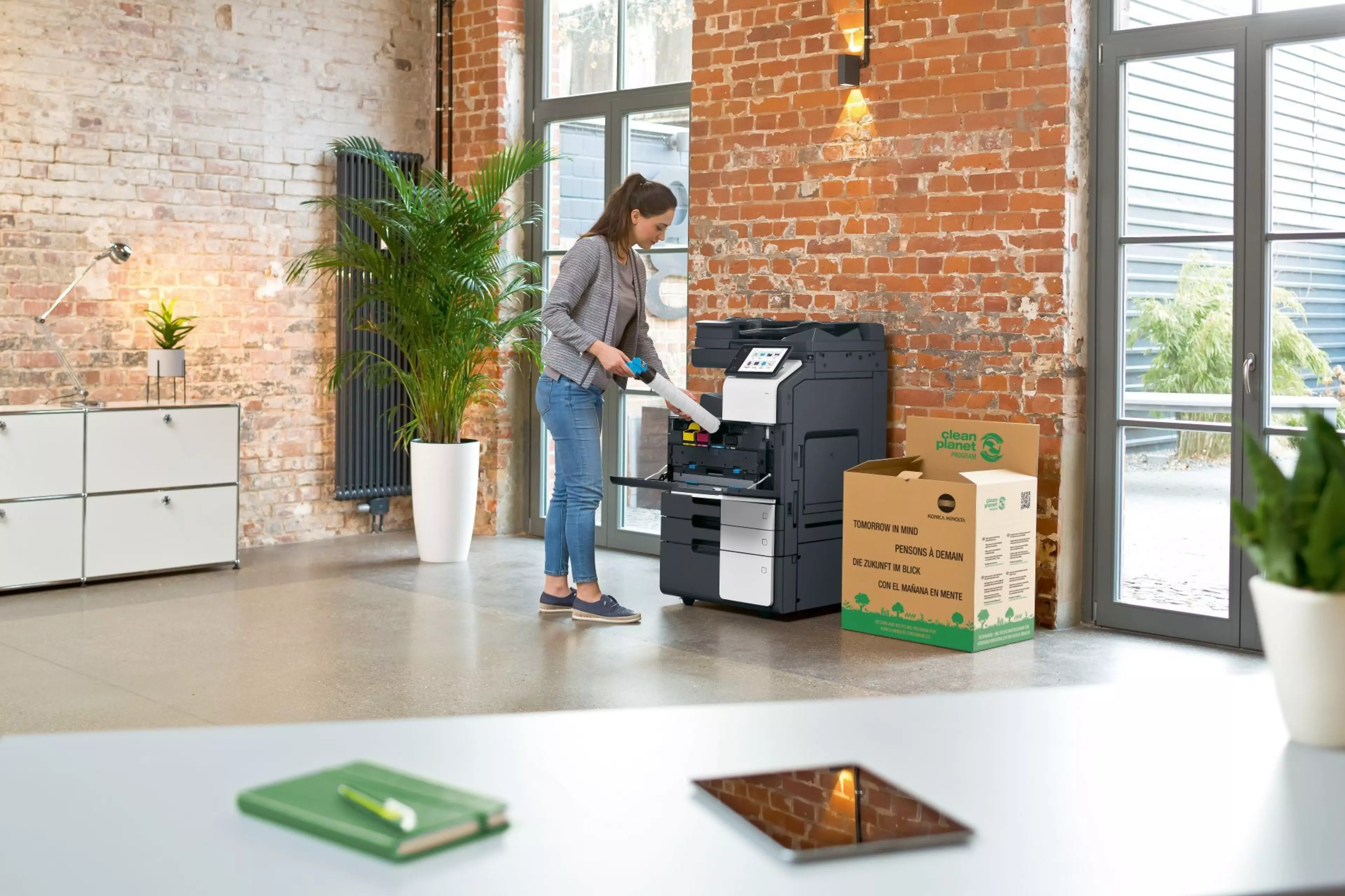 Eine Frau steht in einem modernen Büro und benutzt einen Multifunktionsdrucker von Konica Minolta. Neben dem Gerät steht eine „Clean Planet“-Recyclingbox für umweltfreundliches Tonerrecycling.