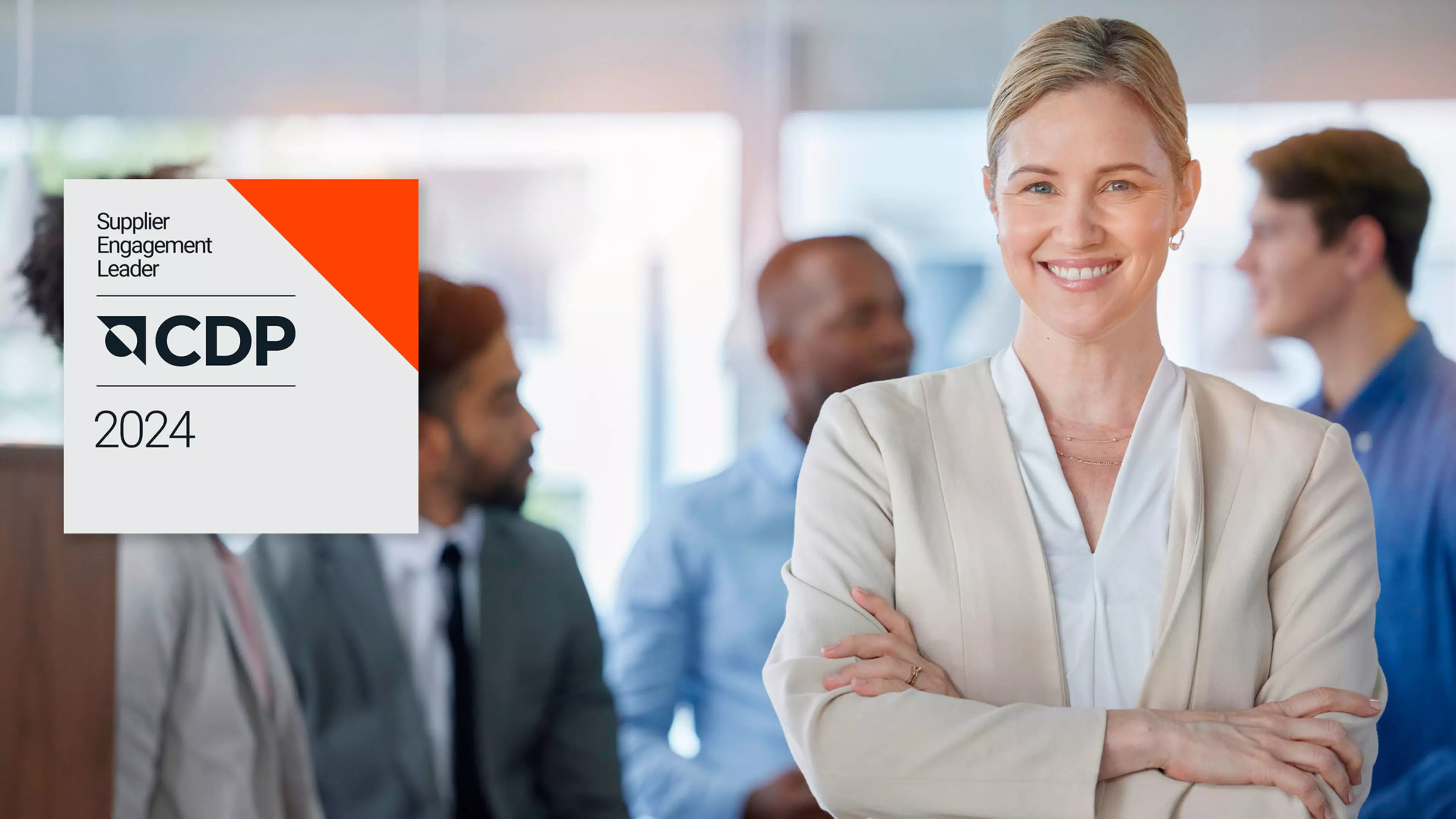 Una mujer joven en una oficina sonríe a la cámara mientras sus compañeros charlan detrás de ella. Sobreimpreso, el logotipo de CDP Supplier Engagement Leader 2024
