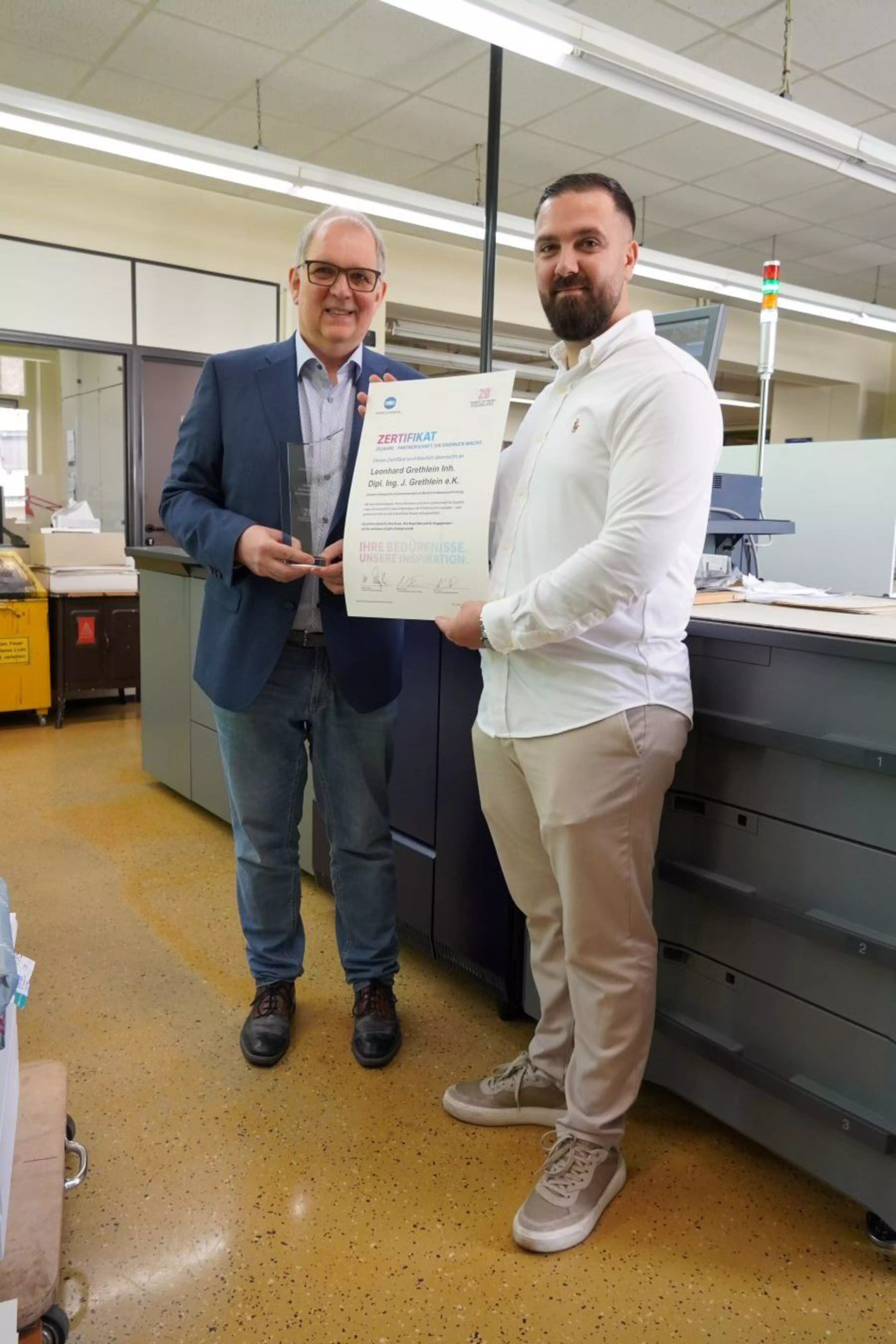 Zwei Personen stehen in einer Druckerei und halten gemeinsam ein Zertifikat, das eine Auszeichnung für den Betrieb zeigt.