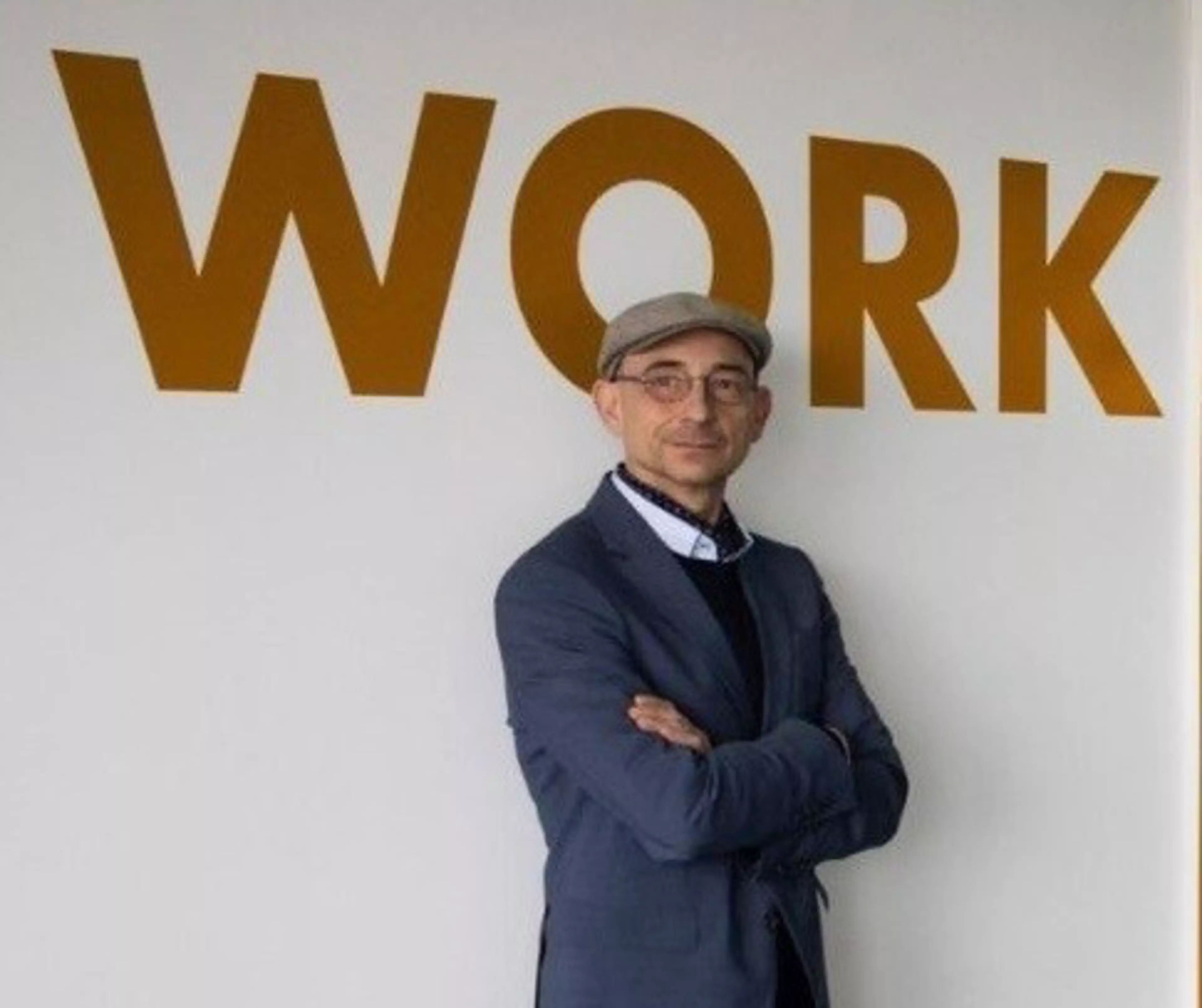 Dr. Martin Weiß steht vor einer Wand mit dem Wort 'WORK' in großen, goldenen Buchstaben und trägt einen Anzug sowie eine Schirmmütze.